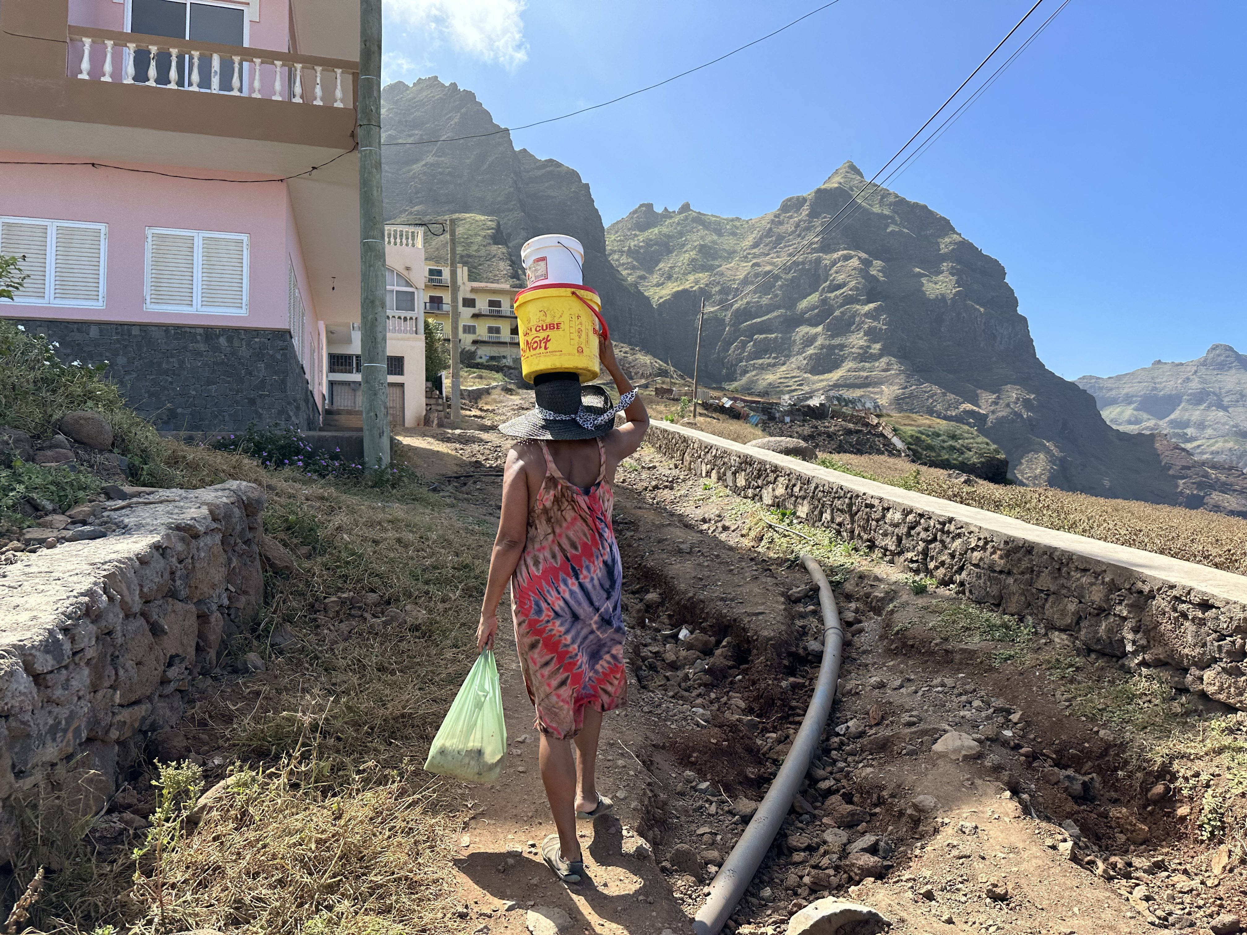 Vrouw met een gele en een witte emmer op het hoofd trekt langs een weg vol kuilen de berg in.